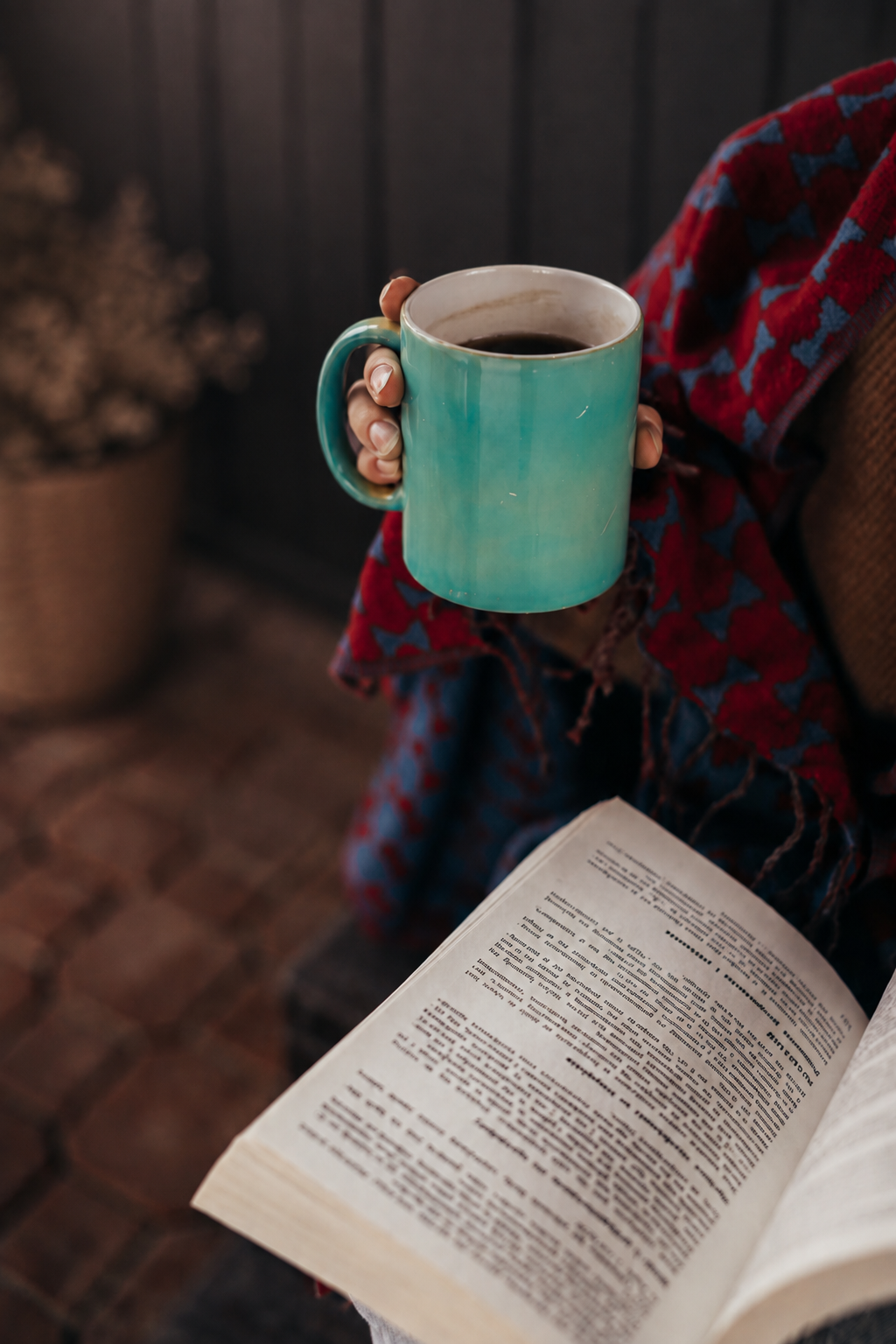 Mãos a segurar uma chávena turquesa sobre um livro aberto, com manta xadrez ao colo