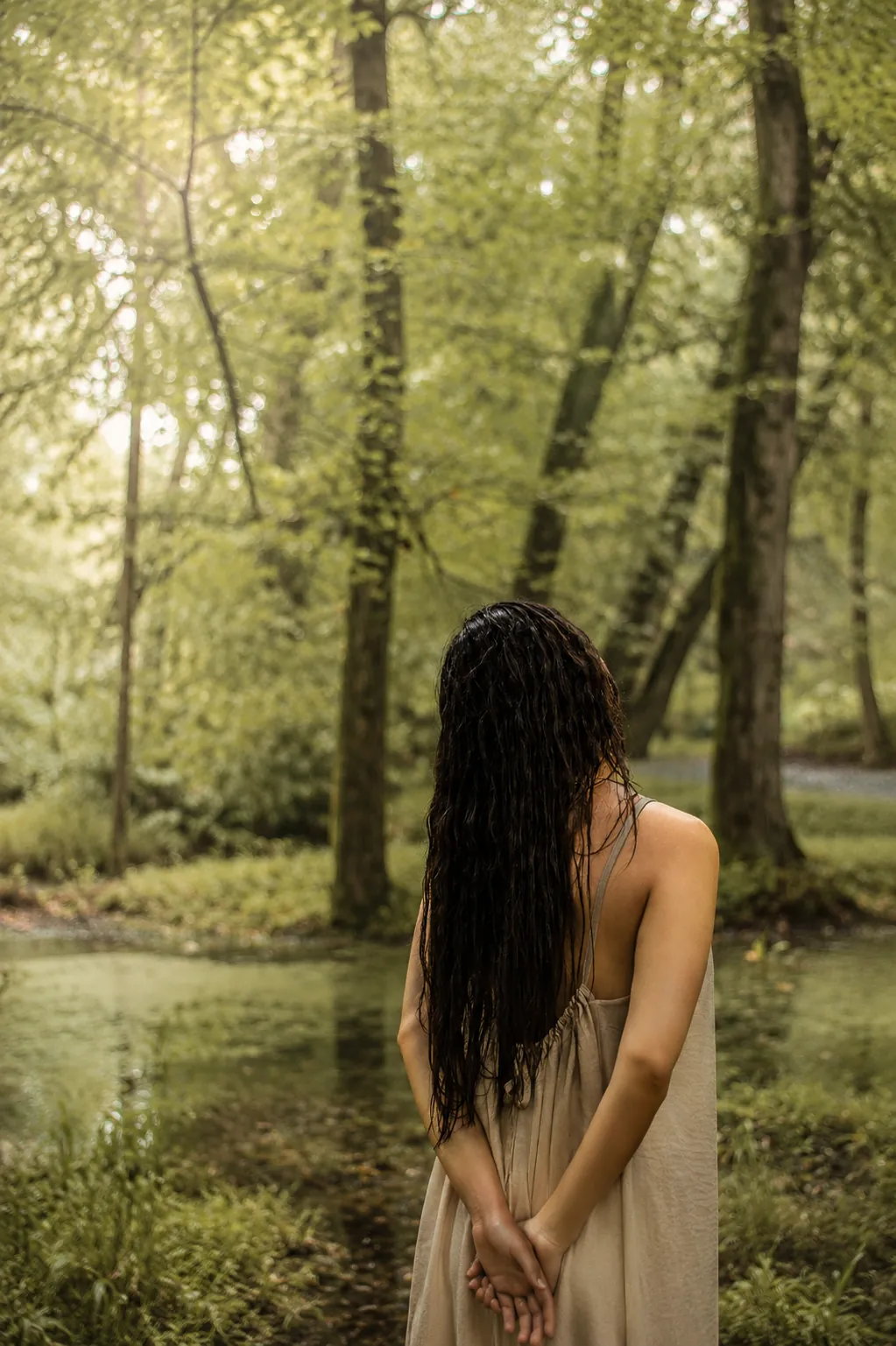 Mulher de costas junto a uma lagoa na floresta, com cabelo escuro e vestido cru, a sentir a água e a vegetação